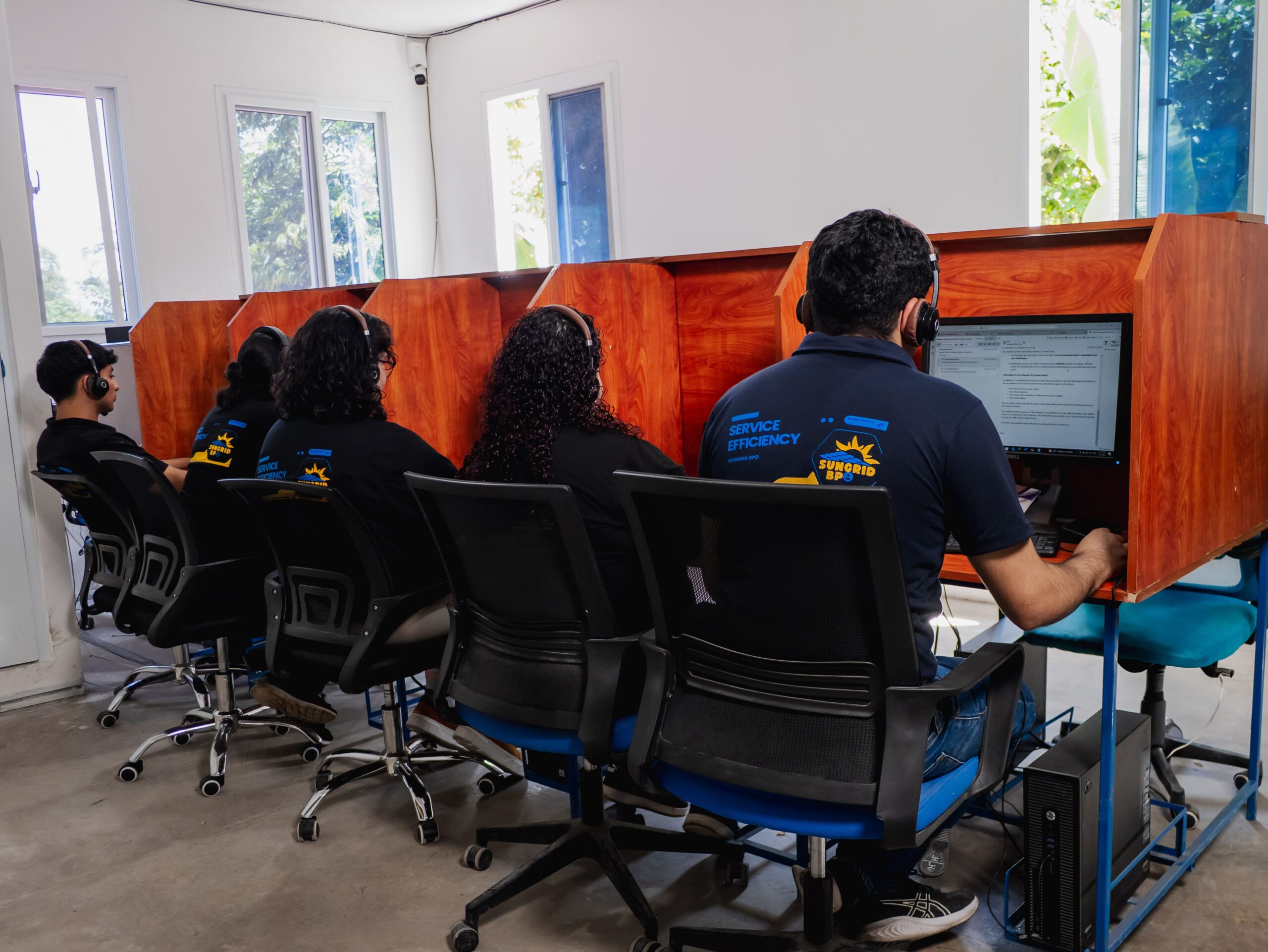 Five people with headsets work at computers in office cubicles, wearing matching company shirts.