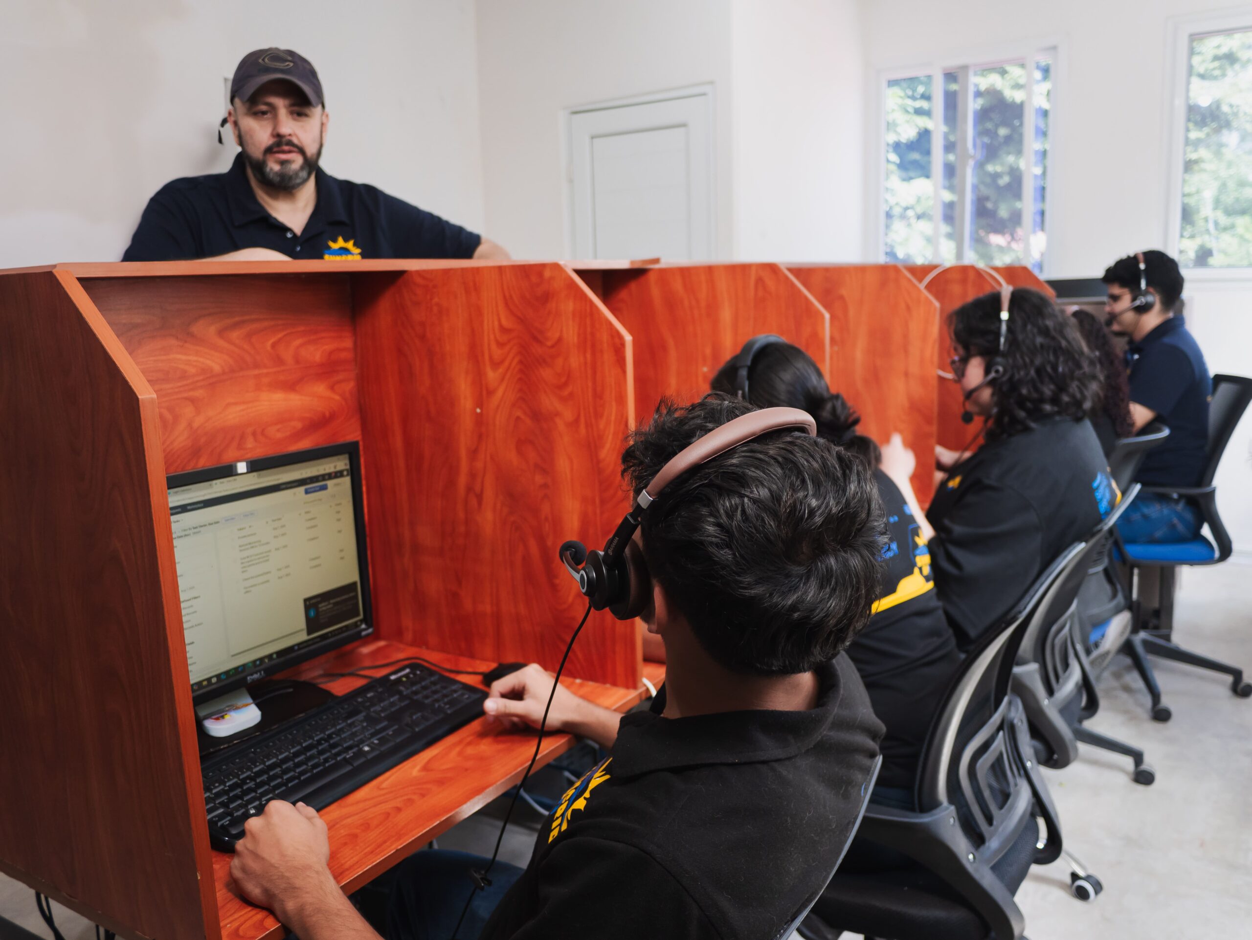 Call center staff with headsets work at computers in wooden cubicles, supervisor stands behind.