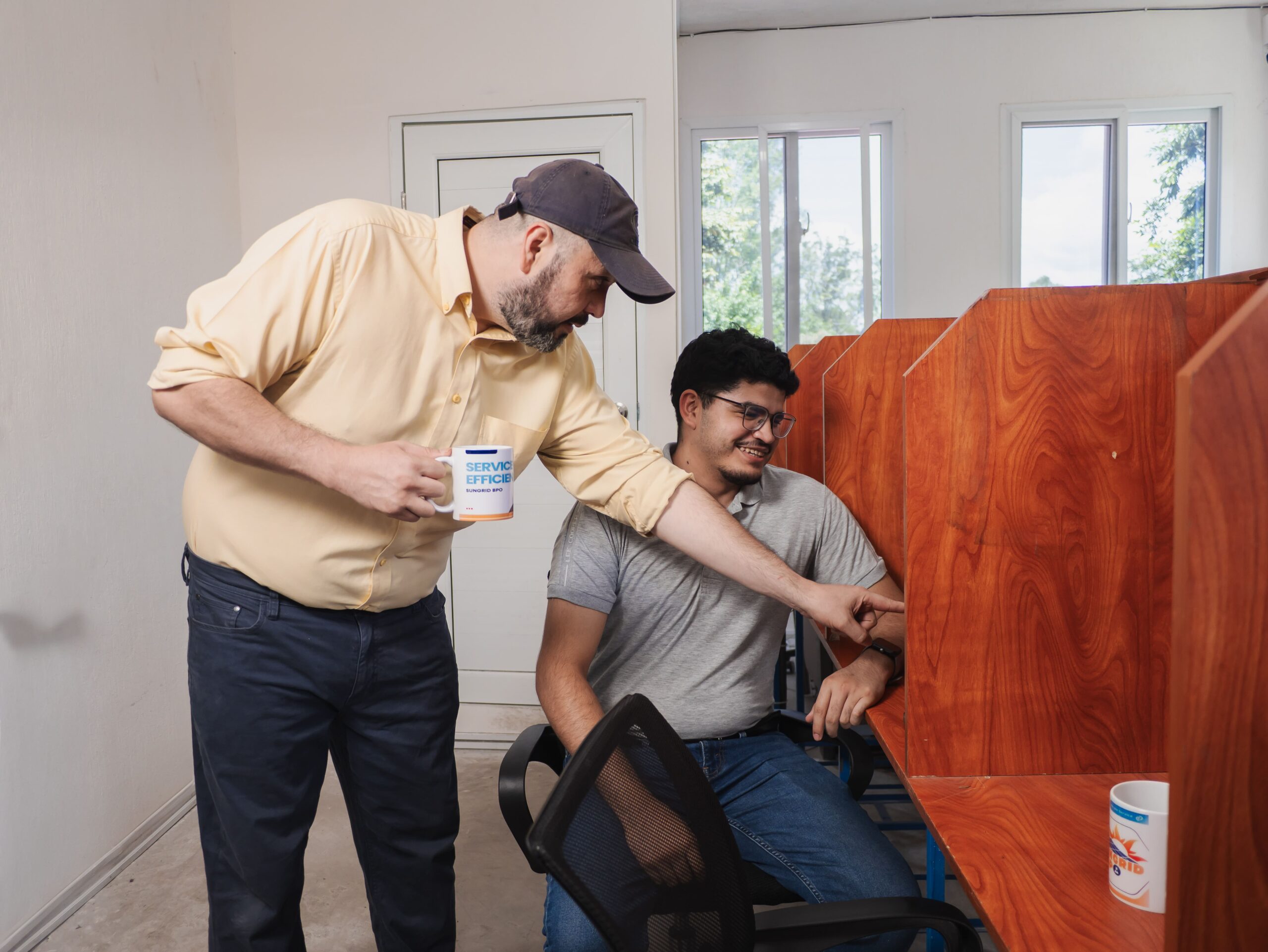 Man in yellow shirt points at something on coworker's desk in office cubicle, holding a "Service Efficiency" mug.